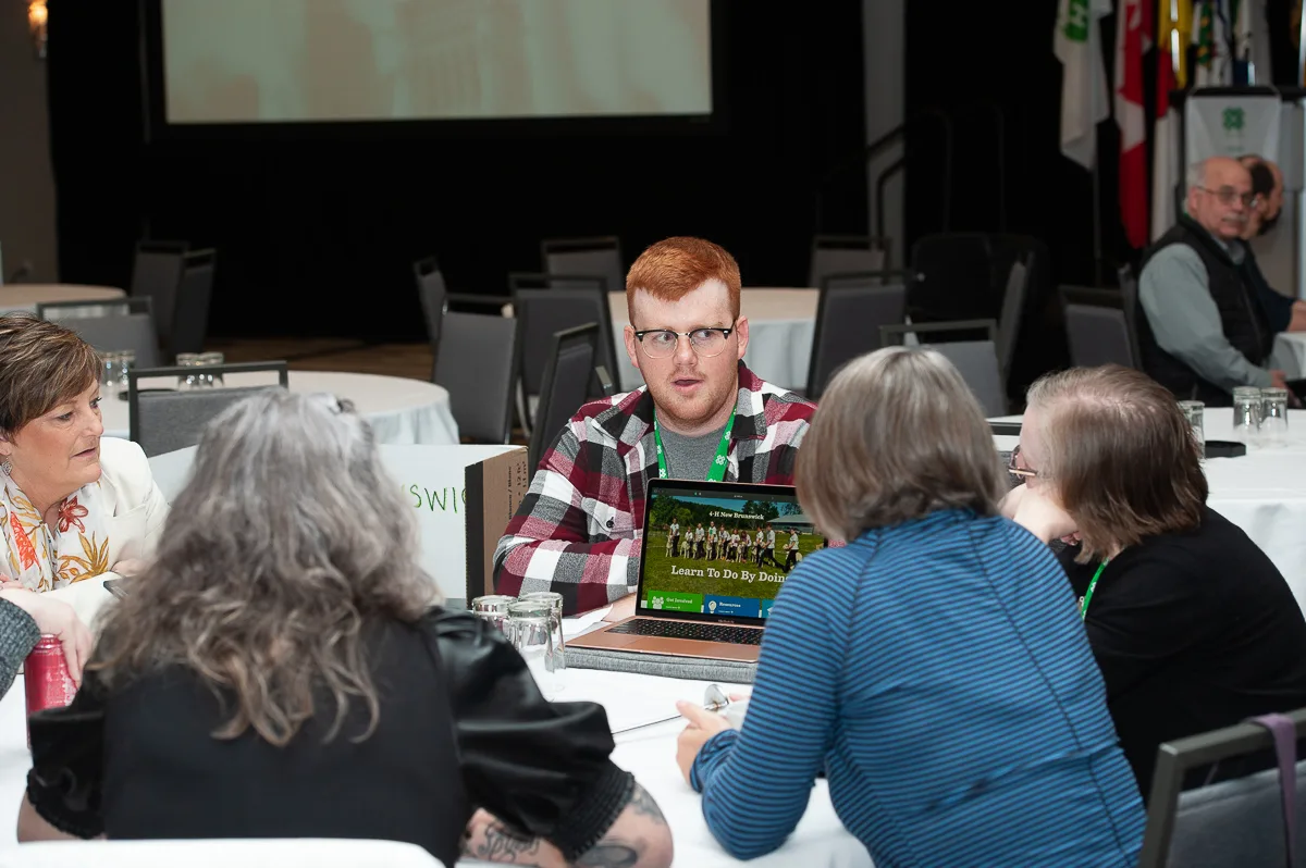 GUn groupe d’adultes assis autour d’une table ronde à une conférence; au centre de la table, on voit un ordinateur portable qui affiche les mots « Apprendre en travaillant »